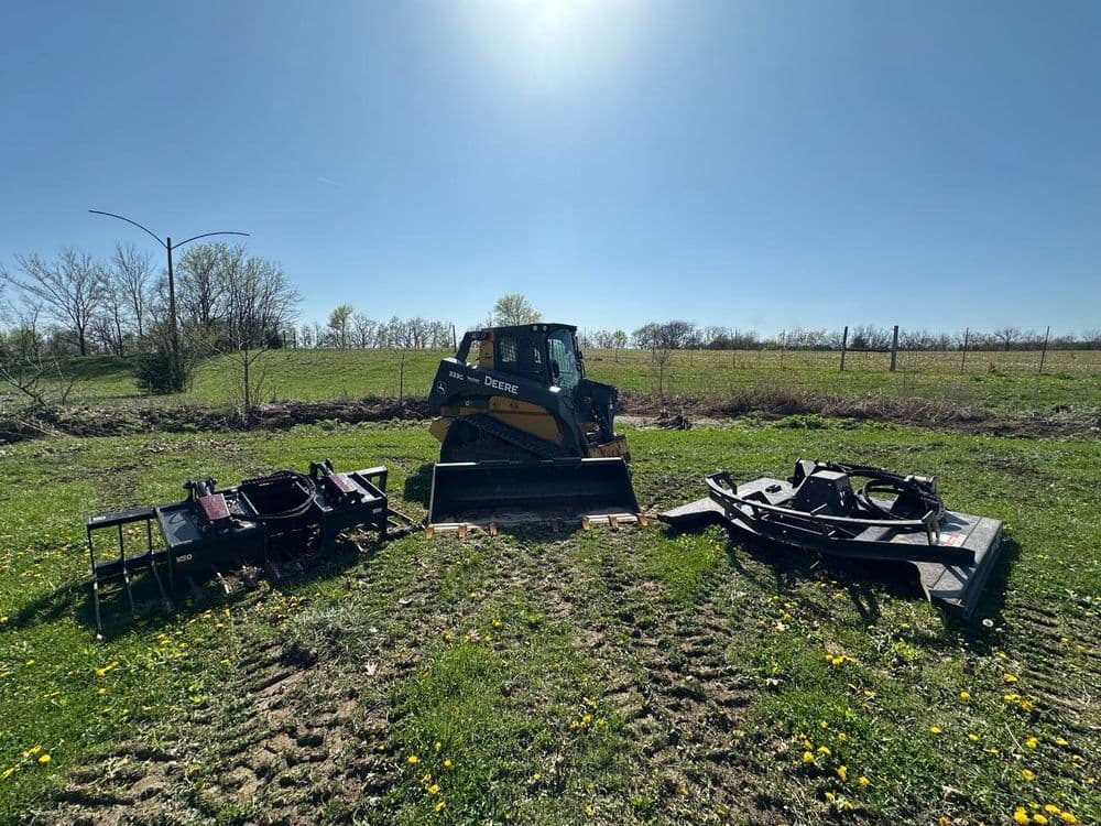 John Deere skid steer loader surrounded by agricultural attachments on a sunny day.