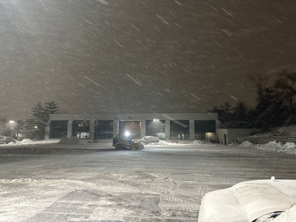 Snowstorm at night with a snowplow clearing a parking lot near a lit building.