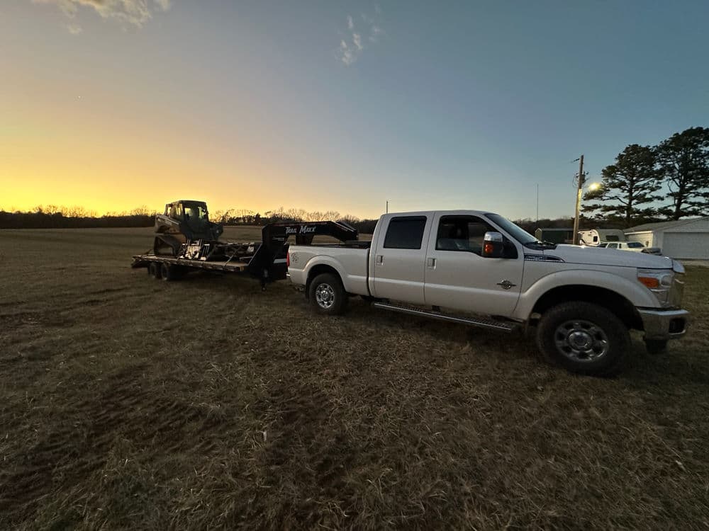 White truck towing a trailer with heavy equipment against a sunset backdrop in an open field.