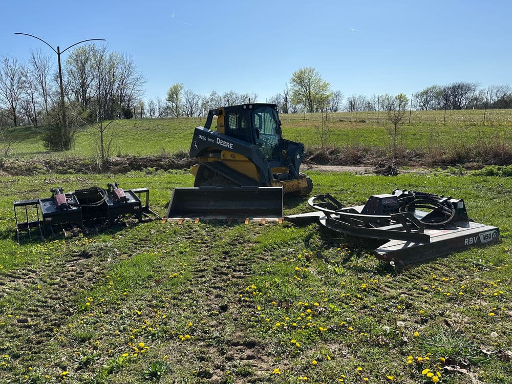 John Deere bulldozer on grassy terrain with agricultural equipment beside it.